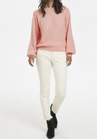 Frau trägt einen rosa gestrickten Pullover mit Pailletten, cremfarbene schmale Hose und schwarze Stiefeletten, während sie vor einem schlichten weißen Hintergrund geht.