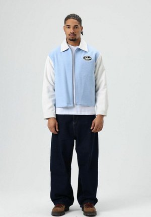 Veste polaire bleu clair avec manches blanches, une fermeture éclair et un logo rond sur la poitrine. Associée à un jean large bleu foncé et des chaussures marron.