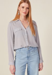 Camisa de manga larga en tela ligera con un patrón geométrico en azul y rojo, que presenta un cuello y dos bolsillos en el pecho, combinada con pantalones de denim claros.