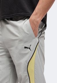 Άνδρας με ανοιχτογκρι παντελόνι Puma με μαύρο λογότυπο και κίτρινη ρίγα, χέρι στην τσέπη, συνδυασμένο με μαύρο πουκάμισο.
