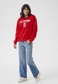Rotes Sweatshirt mit weißem Text, lockere Passform, gerippte Bündchen, kombiniert mit hellblauen weit geschnittenen Jeans und cremefarbenen Sneakers. Einfaches Design, lässiger Look.