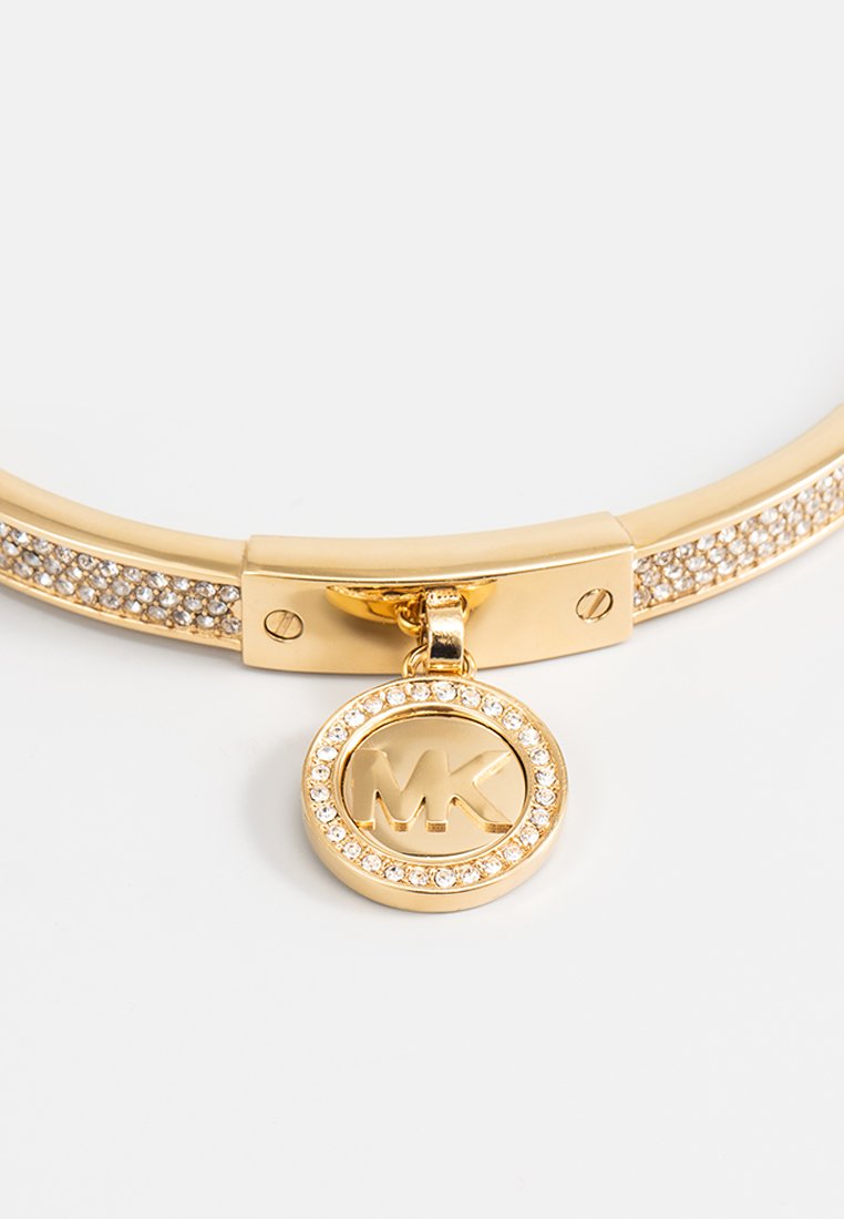 Bracelet doré au design plat, présentant une rangée de cristaux transparents et un pendentif circulaire gravé "MK" entouré de cristaux.