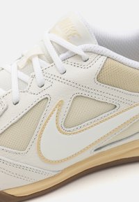 Sapatilha atlética de couro branco com painéis em malha, detalhes em creme e sola de borracha castanha. Apresenta atacadores brancos e uma superfície ligeiramente texturizada.