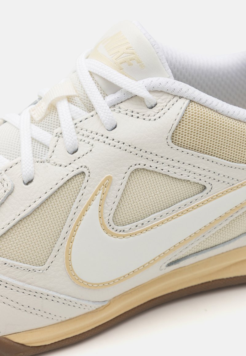 Zapatilla deportiva de cuero blanco con paneles de malla, acentos en crema y suela de goma marrón. Presenta cordones blancos y una superficie ligeramente texturada.