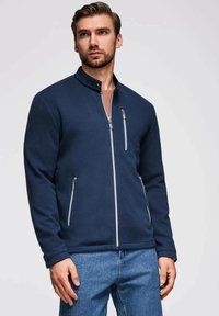 Marinblå zip-up jacka med ribbat mönster, silvrig hårdvara, hög krage och två framfickor. Matchad med ljus denimjeans.