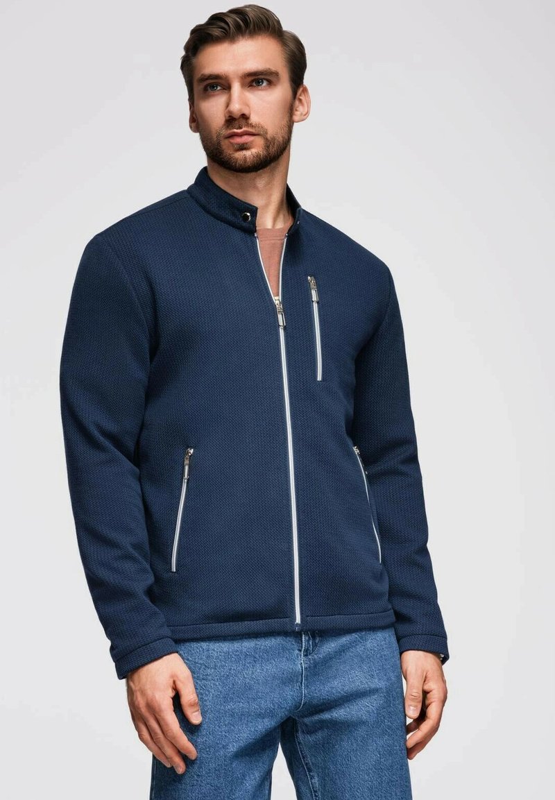 Marinblå zip-up jacka med ribbat mönster, silvrig hårdvara, hög krage och två framfickor. Matchad med ljus denimjeans.