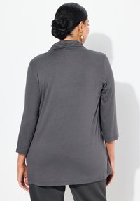Top gris de manga larga con un cuello drapeado y dobladillo redondeado. Tejido suave con una textura lisa. Ajuste modesto, combinado con pantalones oscuros.
