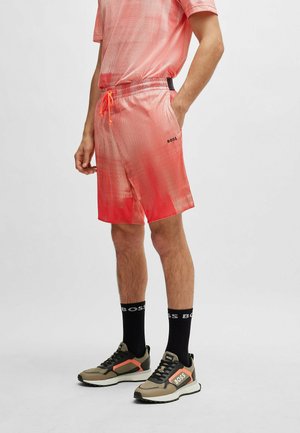 Mann trägt rote Gradient-Shorts und passendes Shirt, schwarze knielange Socken und beige Sneakers mit orangem Detail, Hand in der Tasche.