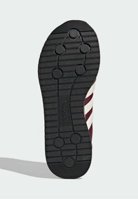 Semelle de chaussure Adidas noire avec motifs circulaires sur la bande de roulement et rainures horizontales, montrant une partie de la tige de chaussure bordeaux avec des bandes blanches.