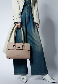 Persona che indossa pantaloni di jeans a gamba larga, stivali bianchi a punta e un trench beige, mentre tiene una borsa in pelle taupe per le maniglie.