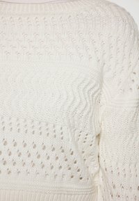Witte gebreide trui met een textuurpatroon, voorzien van golvende lijnen en kleine gaatjes voor ventilatie. Zacht materiaal met een relaxte pasvorm.