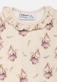 Camisa de color beige claro con un escote con volantes. Presenta un patrón repetido de ardillas de dibujos animados con vestidos sosteniendo flores, adornado con hojas verdes.