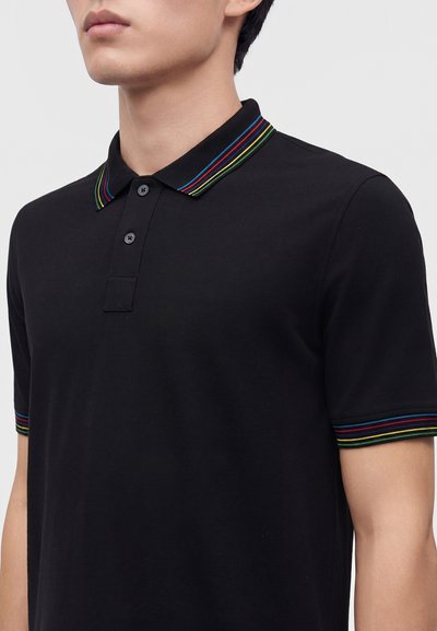 Homme portant un polo noir à manches courtes avec un col présentant des rayures multicolores en rouge, bleu, vert et jaune.
