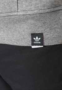 Tessuto di felpa grigia con una texture a coste nella parte inferiore. Etichetta nera con logo bianco adidas in un design a trifoglio. Sfondo nero.