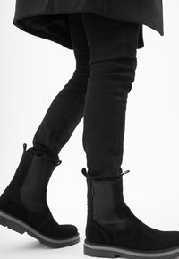 Stivaletti neri in suede con pannelli laterali elastici e linguette per il sollevamento, caratterizzati da una finitura testurizzata e una suola in gomma robusta. Indossati con pantaloni neri attillati.
