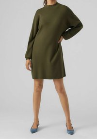 Žena v volném olivově zeleném dlouhém svetrovém šatech a modrých lodičkách s špičatou špičkou, stojící s jednou rukou na boku.