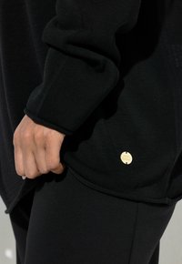 Hand, die den Saum eines schwarzen Langarm-Strickoberteils mit kleinem rundem goldenem Emblem nahe der Taille hält, kombiniert mit schwarzen Hosen.