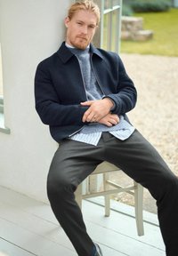Jachetă de bombardier navy peste un pulover tricotat gri, stratificat cu o cămașă cu dungi. Pantaloni închiși, așezat pe un scaun din lemn lângă o fereastră.