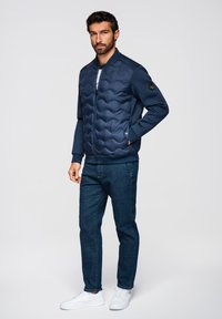 Mand står med den ene hånd i lommen iført en mørkeblå quiltet jakke, mørkeblå jeans og hvide sneakers mod en ensfarvet baggrund.