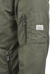 Veste rembourrée verte avec une poche zippée sur le côté et une étiquette portant la mention "BLEND". Le tissu semble lisse avec une finition mate.