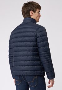 Marineblaue Steppjacke mit hohem Kragen, horizontal gestepptem Design und glatter Textur. Kombiniert mit dunklen Jeans, die einen lässigen Look betonen.