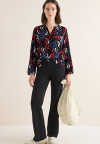 Langärmlige, mehrfarbige Bluse mit roten, blauen und weißen Zickzackmustern, kombiniert mit schwarzen Schlagjeans und einer hellen Tasche.