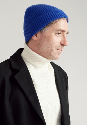 Blauwe ribgebreide beanie met een aansluitend ontwerp, gedragen over een witte kabelgebreide coltrui en een zwarte jas.