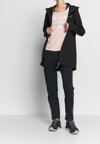 Černá softshellová bunda s kapucí, růžové tričko s výrazným bílým textem a námořnické tepláky s bočními akcenty. Celkový vzhled doplňují černé sportovní boty.