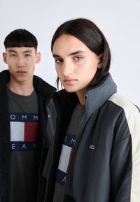 Chaqueta gris con cremallera y mangas en crema contrastantes, llevada sobre una camiseta gris con un logotipo rojo y azul. Tejido suave y texturizado.