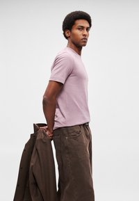 Νεαρός άνδρας με κοντά σγουρά μαλλιά, φορώντας ροζ μπλουζάκι και καφέ παντελόνι, κρατώντας ένα καφέ σακάκι πίσω από την πλάτη του, με απλό φόντο.