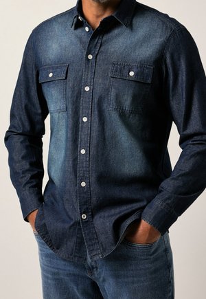 Homme portant une chemise en denim bleu foncé à boutons avec deux poches poitrine et un jean bleu, mains dans les poches, sur fond uni.