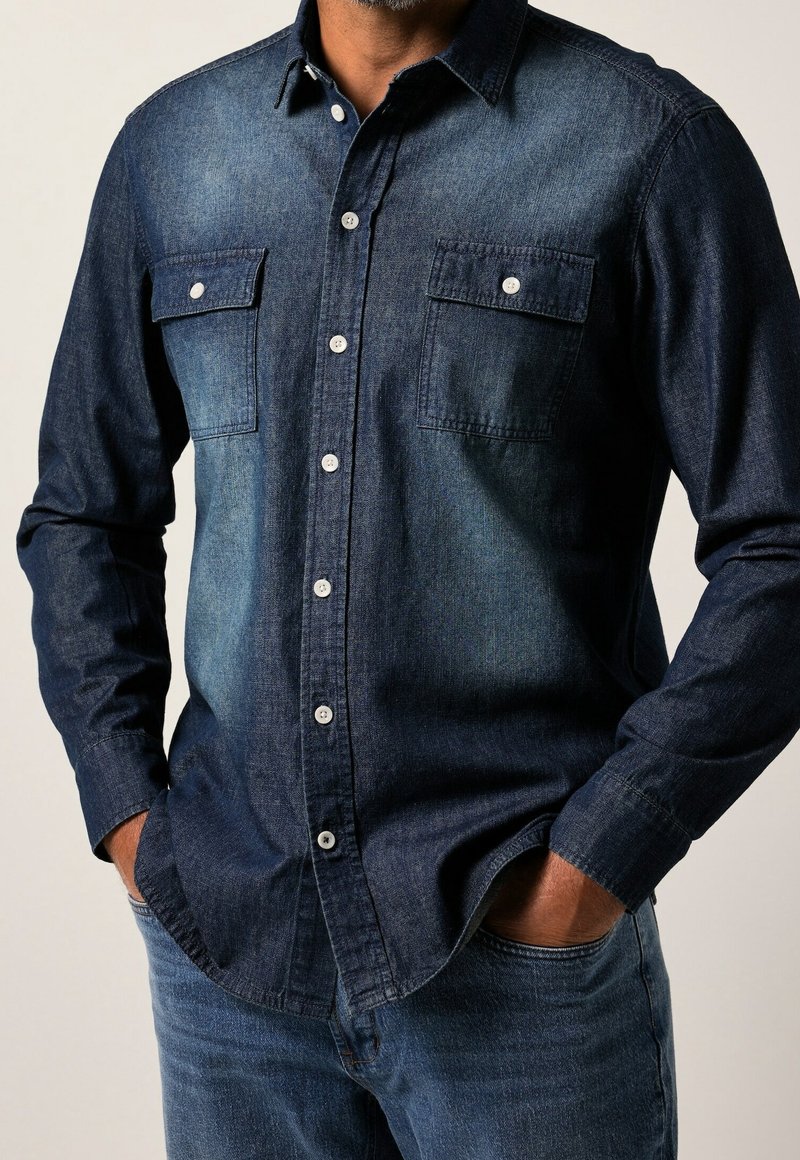 Homme portant une chemise en denim bleu foncé à boutons avec deux poches poitrine et un jean bleu, mains dans les poches, sur fond uni.