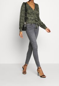Top envolvente de leopardo verde con mangas largas, combinado con jeans skinny grises y tacones altos negros con tiras, acentuado por un pequeño bolso negro.