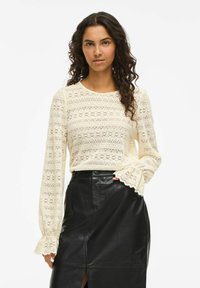 Vrouw met krullend donker haar, gekleed in een crèmekleurige kanten blouse met lange mouwen en een zwarte leren rok, staat met één hand in haar zak.