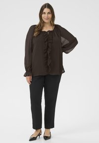 Mujer de pie con blusa de manga larga marrón oscuro con volante en el frente, pantalones ajustados negros y tacones de slingback negros con punta en un fondo blanco.