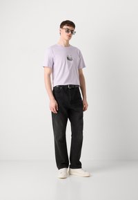 Lilac cotton t-shirt with a graphic print, paired with black denim pants and white sneakers. Accessories include gray sunglasses.