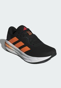 Zwarte sportschoen met bovenwerk van mesh, voorzien van drie oranje strepen, rubberen zool en witte tussenzool. Geaccentueerd met een rood logo op de tong.