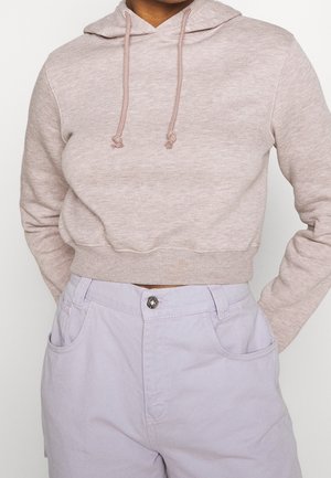 Persona con una felpa corta beige chiaro con coulisse e pantaloni lavanda chiaro a vita alta, mani appoggiate dietro la schiena.