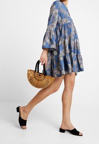 Modré šaty s vzorem a zvonovými rukávy, vrstvený design, černé semišové muly a kabelka ve tvaru půlměsíce z bambusu s černými uchy.