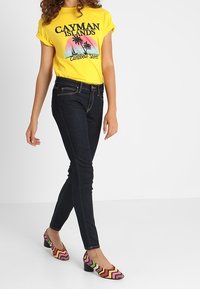 Camiseta amarilla de manga corta con texto gráfico negro, combinada con jeans skinny oscuros y zapatos de tacón bloque con un patrón colorido.