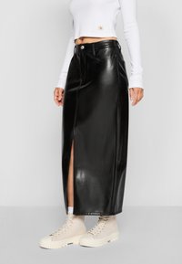 Zwarte faux leren maxi-rok met een split aan de voorkant, voorzien van een hoge taille en knoopsluiting, gecombineerd met lichtgekleurde veterschoenen.