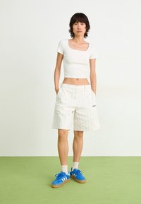 Bílý vypasovaný crop top, bílé šortky s jemným proužkem a kapsami, modré semišové tenisky s bílými pruhy, stojící na zeleném koberci.