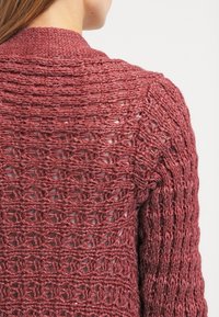 Gilet au crochet de couleur bordeaux, présentant un motif à mailles ouvertes avec des détails texturés et une coupe décontractée, mettant en valeur le décolleté et les épaules.