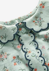 Tissu vert doux avec de petits motifs floraux, bords festonnés bordés de bleu marine, et deux boutons blancs sur l'encolure d'un vêtement.