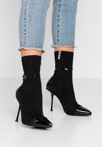 Botines de tacón stiletto negros con un upper de tela ajustado y puntera de charol en forma de punta. Cuenta con un cierre de cremallera y un detalle decorativo de correa.