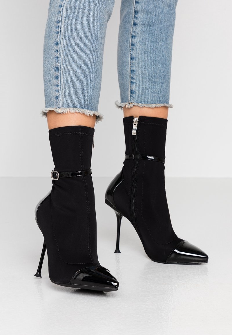 Botines de tacón stiletto negros con un upper de tela ajustado y puntera de charol en forma de punta. Cuenta con un cierre de cremallera y un detalle decorativo de correa.