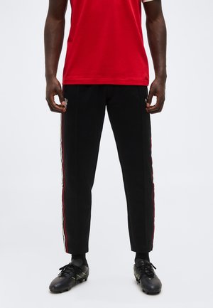 Άνδρας που φοράει μαύρο αθλητικό παντελόνι με κόκκινες και λευκές λωρίδες στο πλάι, μαύρα ποδοσφαιρικά παπούτσια και κόκκινο αθλητικό πουκάμισο, στέκεται σε λευκό φόντο.