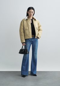 Padded beige jacket with shearling collar, black top, flared blue denim jeans, black handbag, and pointed toe black shoes.