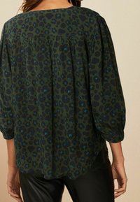Blouse à imprimé léopard vert avec des manches trois-quarts, des plis au dos et un col arrondi. Texture de tissu douce.