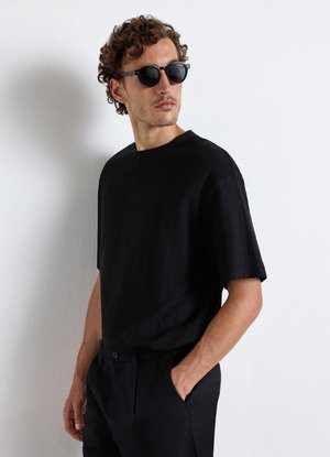 Mann mit lockigem Haar, der schwarze Sonnenbrille, ein schwarzes Kurzarmshirt und schwarze Hose trägt, steht mit einer Hand in der Tasche vor einer weißen Wand.